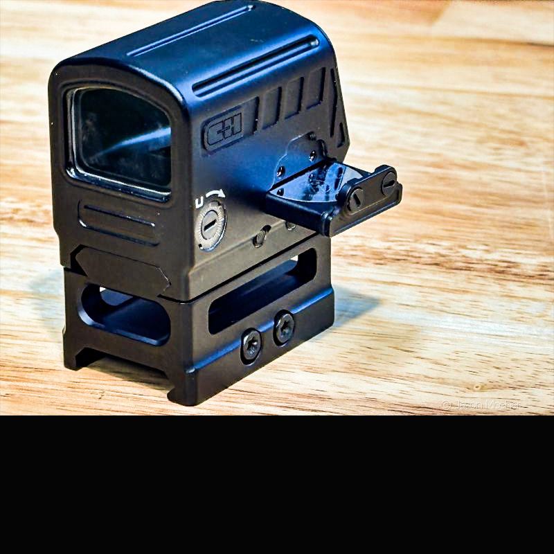 A black red dot sight on a light wood surface with the small battery trey pulled open. 