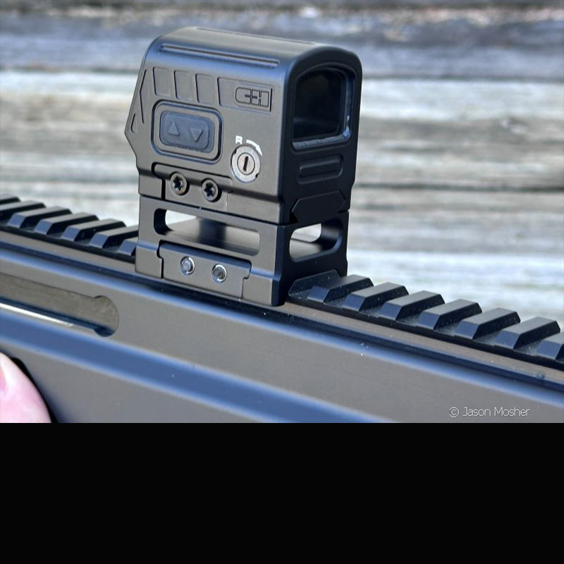 A black optic mounted to the rail of a gun, displayed Infront of a grey wood background. 
