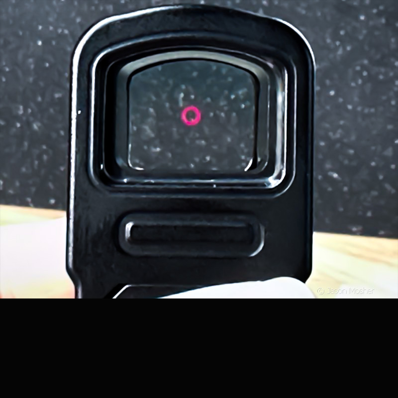 Looking through the lens of a black optic to see a red circle reticle. 