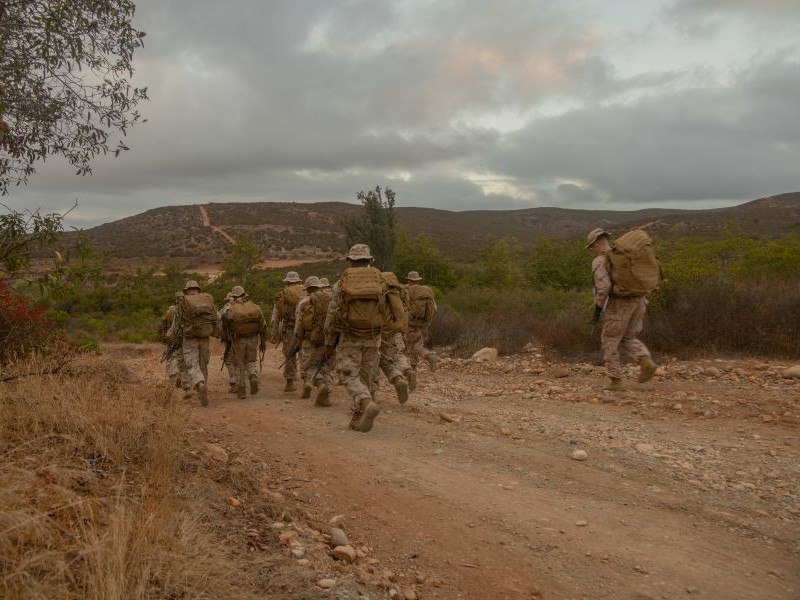 A group o marines with military gear marching down a dirt path with hills in the background. 