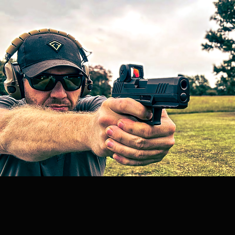 A person wearing a hat, sunglasses, and a black cap, shooting a handgun with the Vortex Defender CCW mounted on it. 
