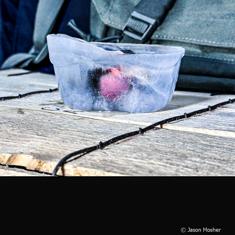 The CCW Defender Solar enclosed optic frozen in a cube of ice, setting on a wooden box. 
