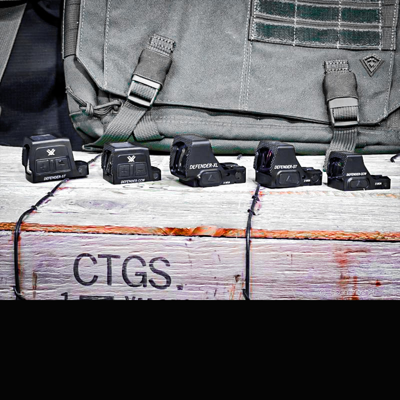 All of the Vortex handgun optics in the Defender series, lined up on a wood box with a green background.