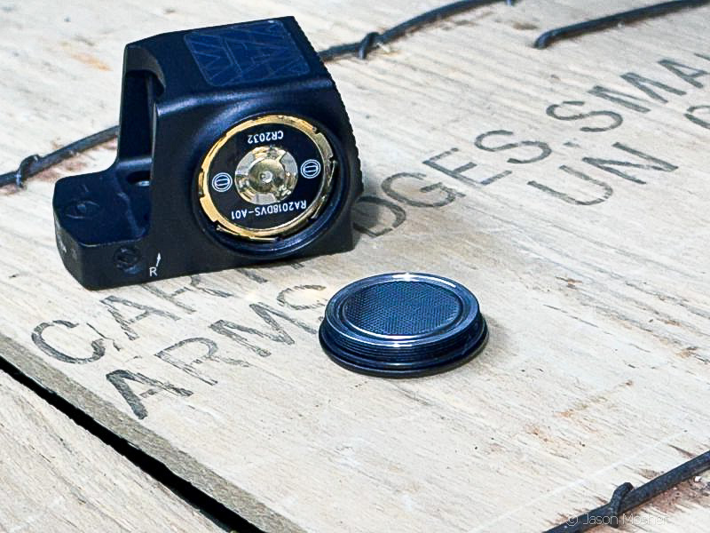 The Defender-ST optic with on a wood box with the battery cap and battery laying beside it. 