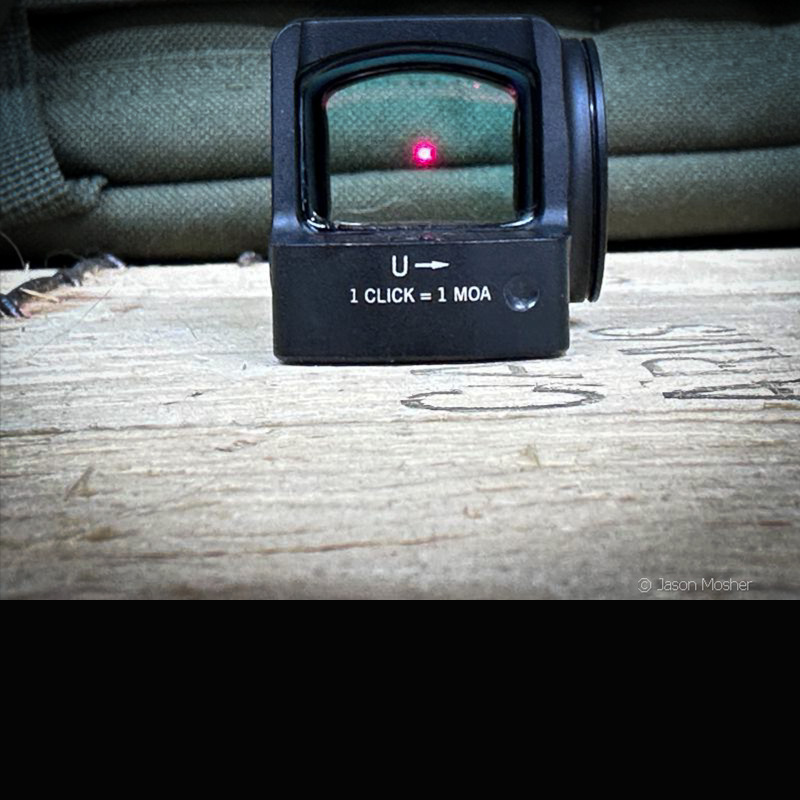 A view of the back of the Venom red dot, with the red dot on. The dot is visible through the glass, against a green bag. 