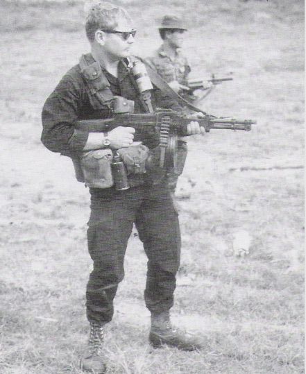 A black-and-white photo shows a MACV-SOG soldier with sunglasses and a chest rig. He is holding a modified, "chopped" RPD light machine gun with a drum magazine, standing in a field with another soldier behind him.