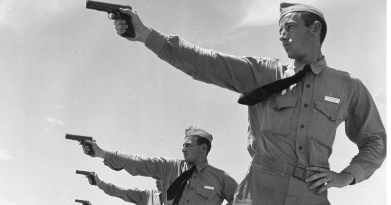 A black-and-white photo shows a group of men in a line, dressed in vintage military uniforms, practicing pistol shooting.