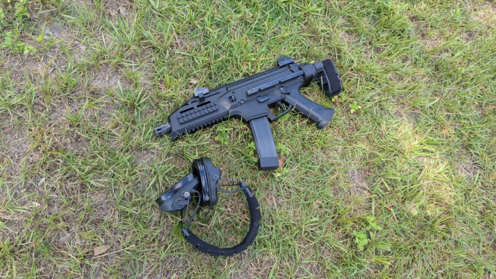 A black CZ Scorpion Evo 3 pistol with a brace lies on the grass next to a pair of black hearing protection headphones.