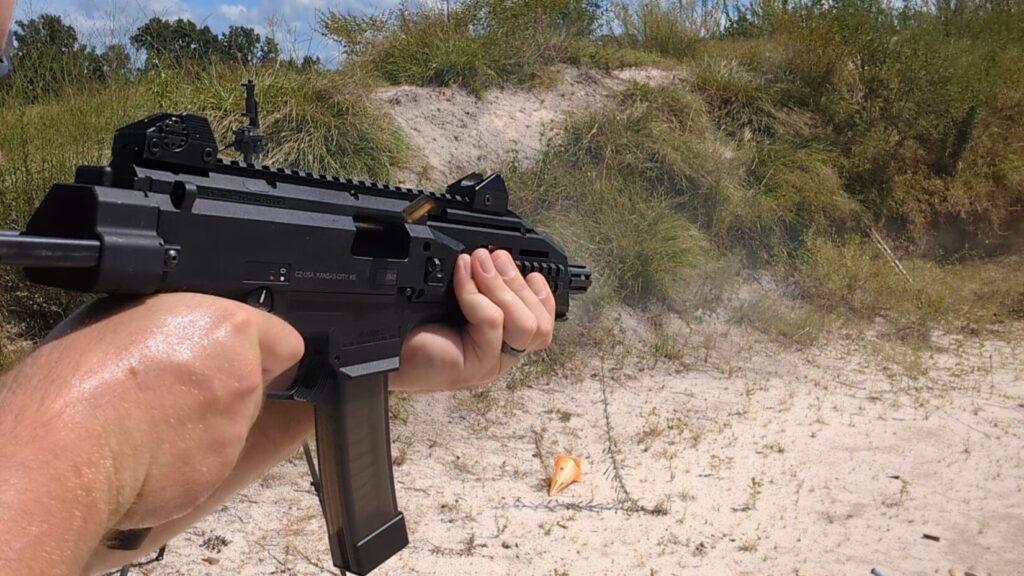  A man with a beard, sunglasses, and hearing protection aims a black CZ Scorpion Evo 3 pistol at a firing range.