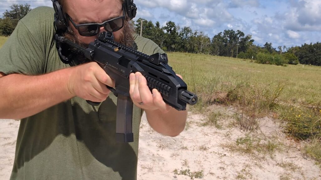 A man with a beard and sunglasses fires a black CZ Scorpion Evo 3 pistol, with a spent brass casing visible in the air.