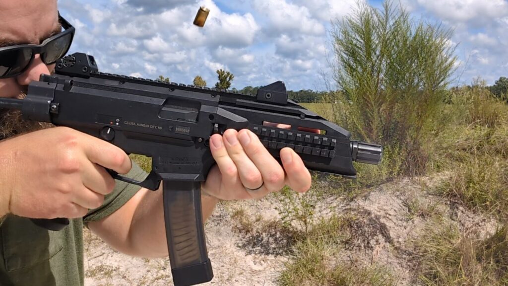 A man with a beard and hearing protection fires a black CZ Scorpion Evo 3 pistol, with a brass casing ejecting from the weapon's action.