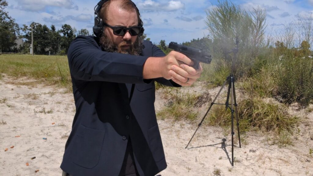 A man with a beard and sunglasses, wearing a black blazer, fires a pistol. The muzzle flash is visible as he shoots.