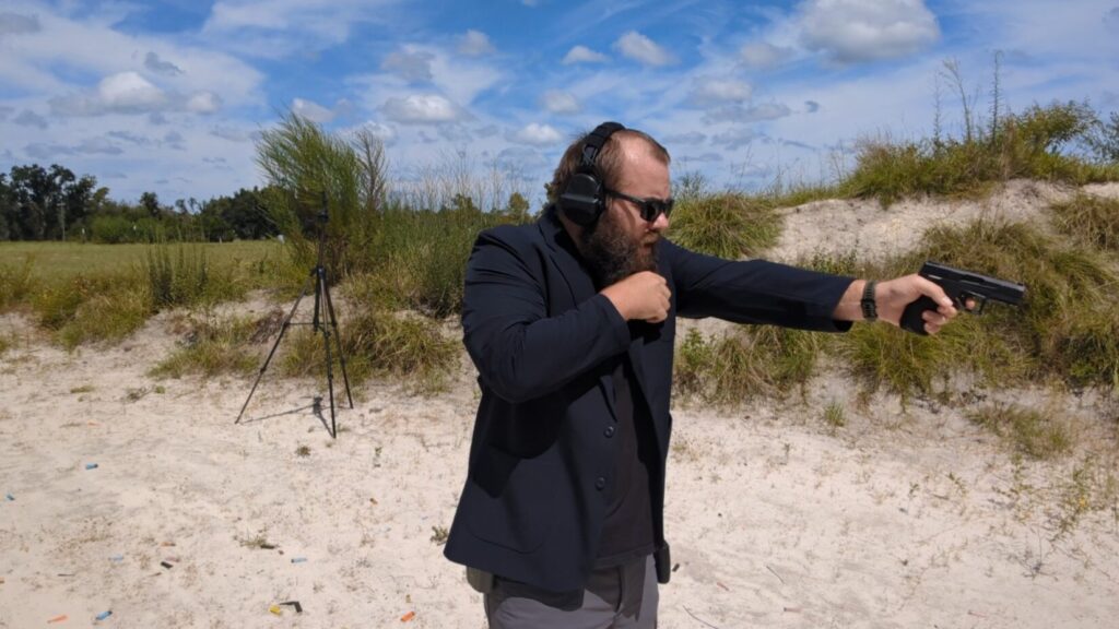 A man with a beard and sunglasses, wearing a blazer, aims a pistol at a firing range while pulling back his opposite arm.