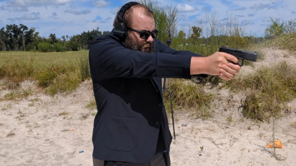 A man with a beard and sunglasses shoots a pistol, with smoke and an ejected shell casing visible.