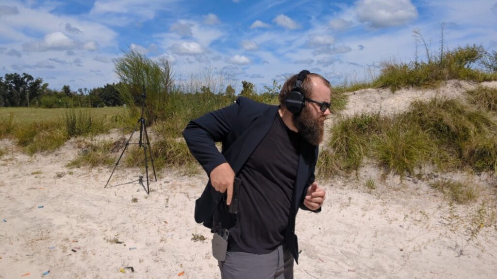  A man with a beard and sunglasses draws a pistol from a holster concealed by his blazer at an outdoor range.