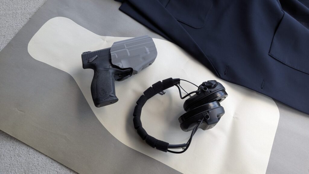 A close-up of a handgun in a holster and ear protection headphones resting on a paper shooting target next to a blue jacket.