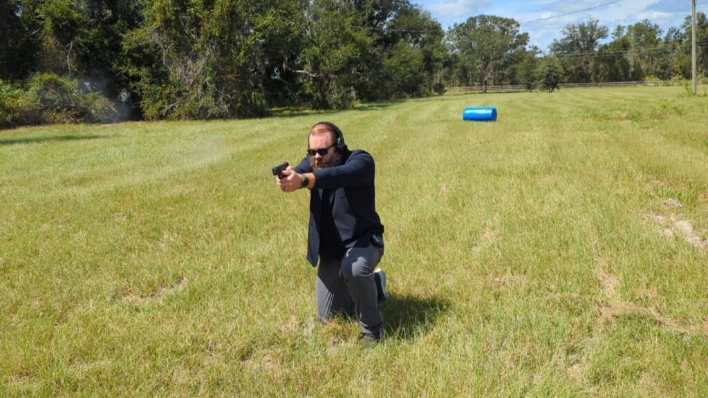 A man with a beard and sunglasses, wearing a black blazer and grey pants, kneels at a firing range aiming a pistol.