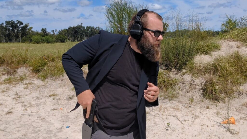A man with a beard and sunglasses draws a pistol from a concealed holster under his blazer at a firing range.
