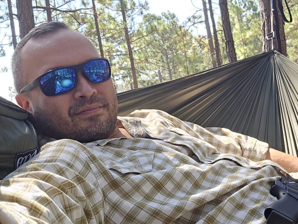 A man in a plaid shirt with sunglasses on rests in a hammock as a chair.