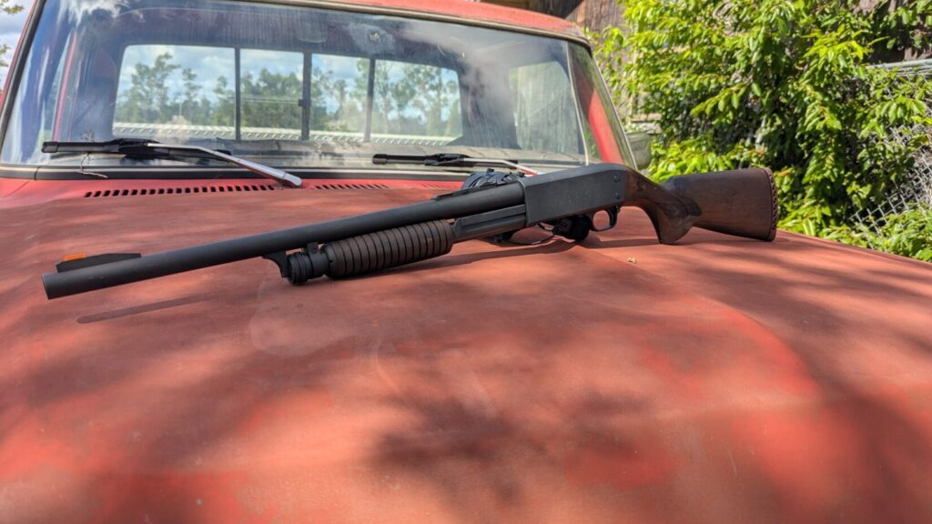 A black shotgun with a wooden stock rests on the rusty hood of a red pickup truck.