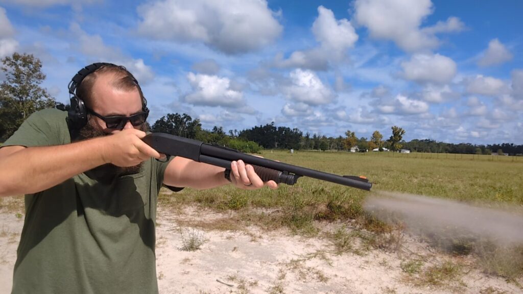 A man with a beard and sunglasses holds a shotgun at a firing range