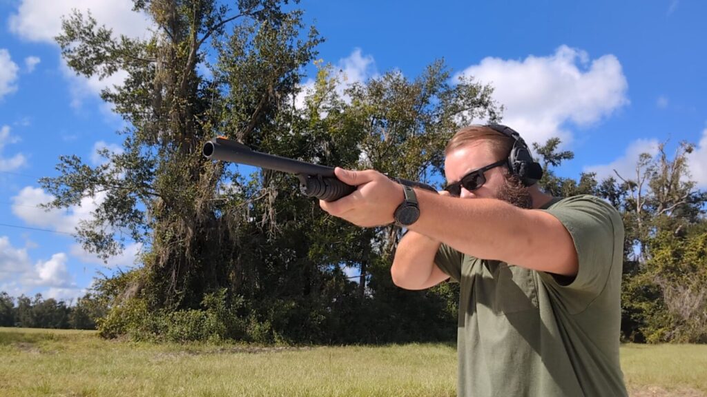 A man with a beard and sunglasses aims a shotgun, with his face mostly obscured by the gun