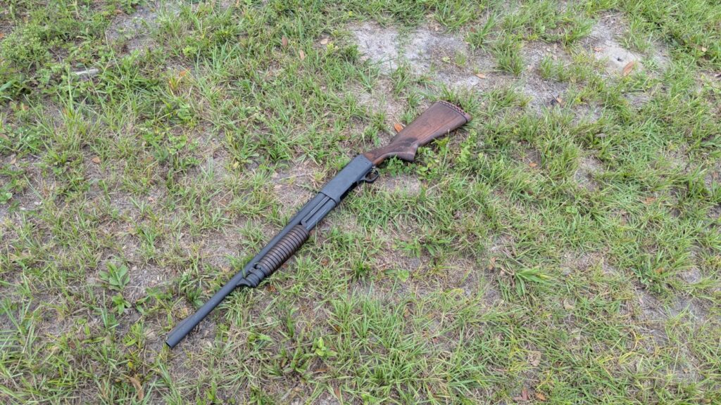 A dark-colored shotgun with a wooden stock rests in a grassy field.