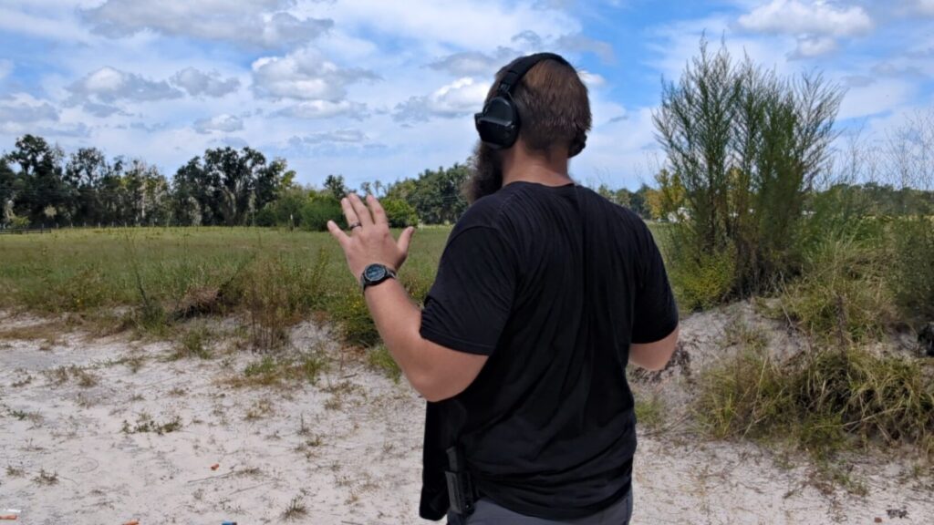 A man with a beard and hearing protection, wearing a black t-shirt, stands at a firing range with his back to the camera, raising a hand.