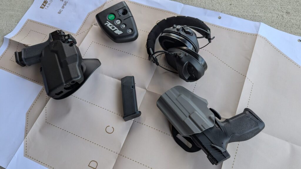 two guns, holsters, ear protection, and a timer are laid out on a paper shooting target.