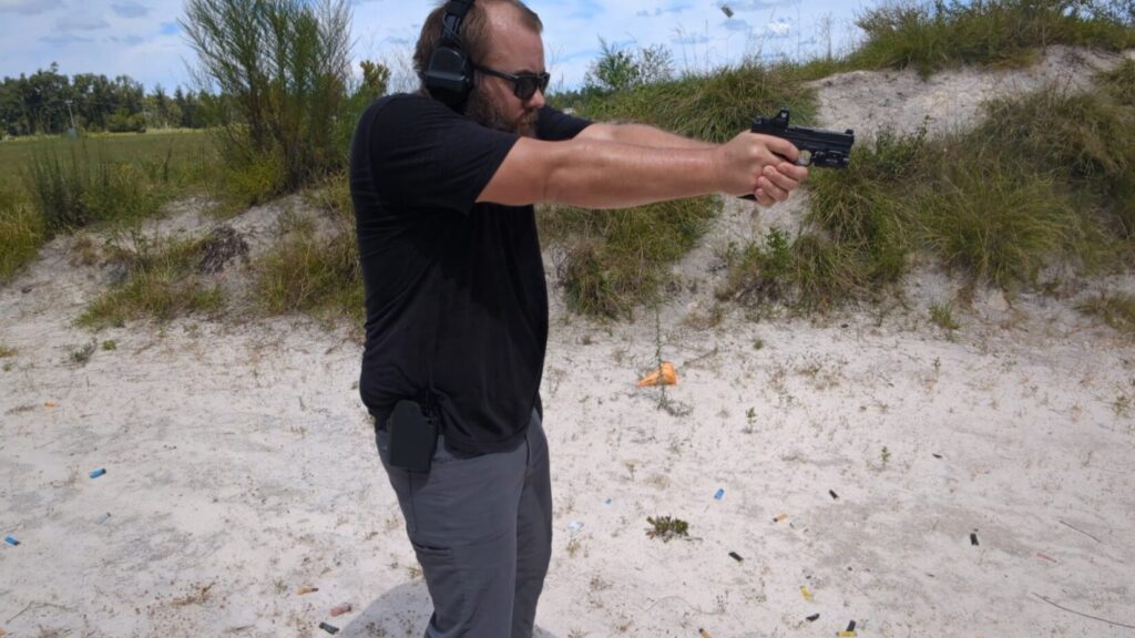A man with a beard and sunglasses shoots a pistol at an outdoor range, with two ejected shell casings visible in the air.
