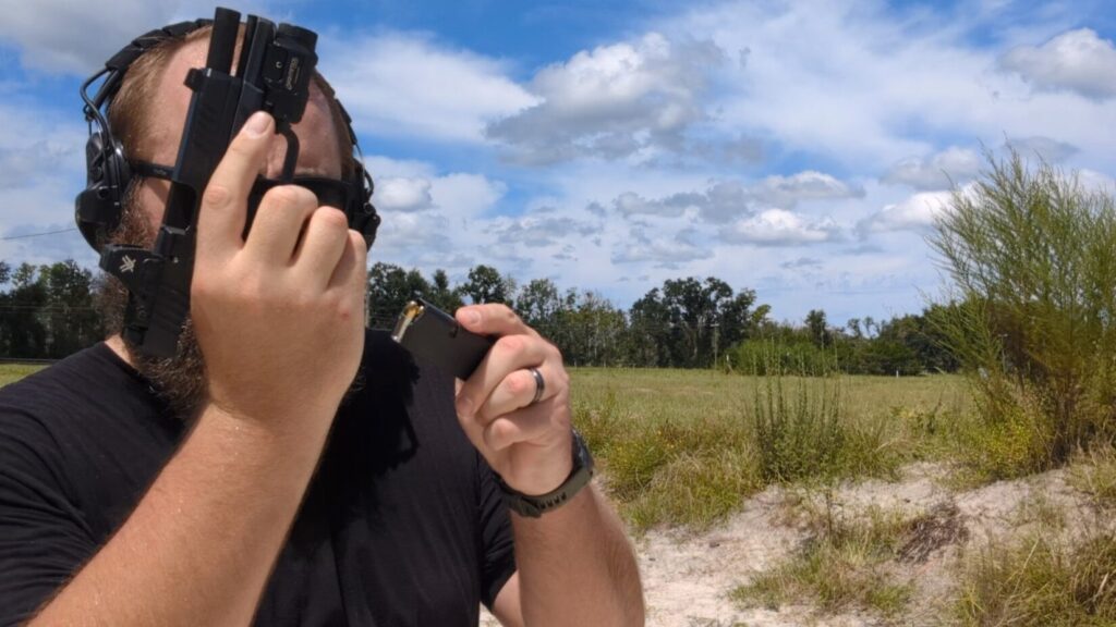 A man with a beard and sunglasses performs a tactical reload, inserting a new magazine into his pistol while dropping the old one.