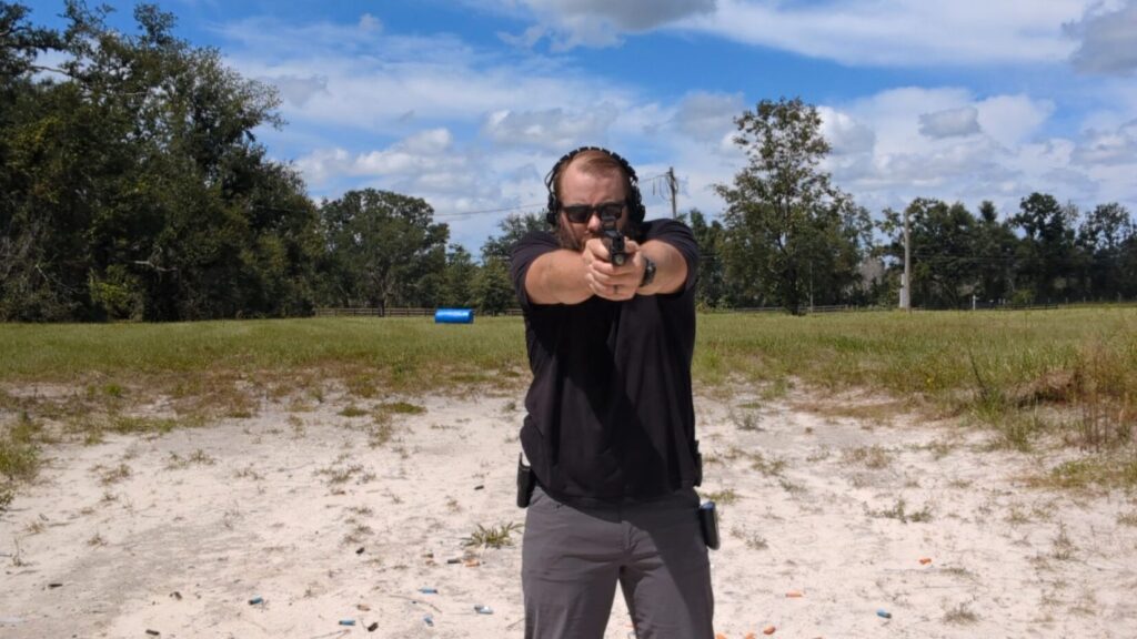 A man with a beard and sunglasses aims a pistol with two hands at the camera.