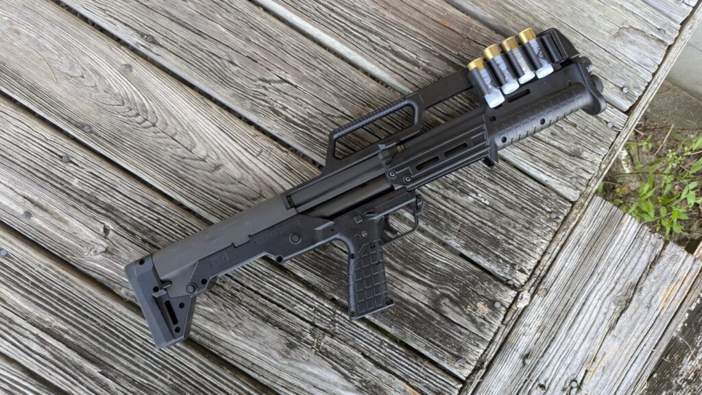 A black KelTec KS7 shotgun, with shells visible in its carry handle, is placed on a worn wooden deck.