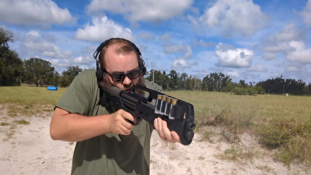 A man with a beard, sunglasses, and hearing protection aims a KelTec KS7 shotgun at a firing range.