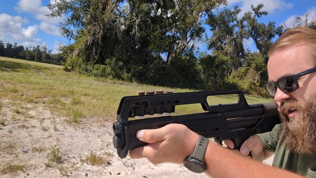 A man with a beard, sunglasses, and hearing protection aims a KelTec KS7 shotgun from the side at a firing range.