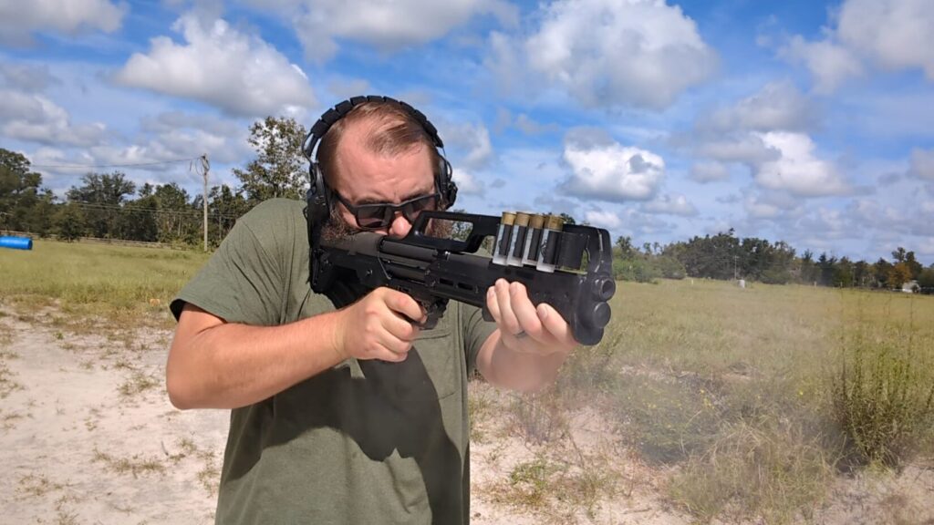 A man with a beard and sunglasses, wearing hearing protection, aims a KelTec KS7 shotgun at an outdoor range.