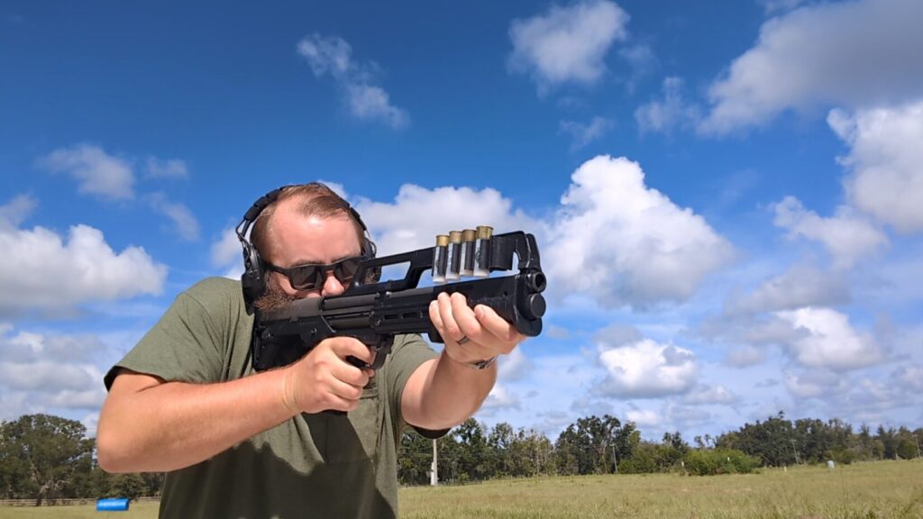 A man with a beard and sunglasses, wearing hearing protection, fires a KelTec KS7 shotgun.