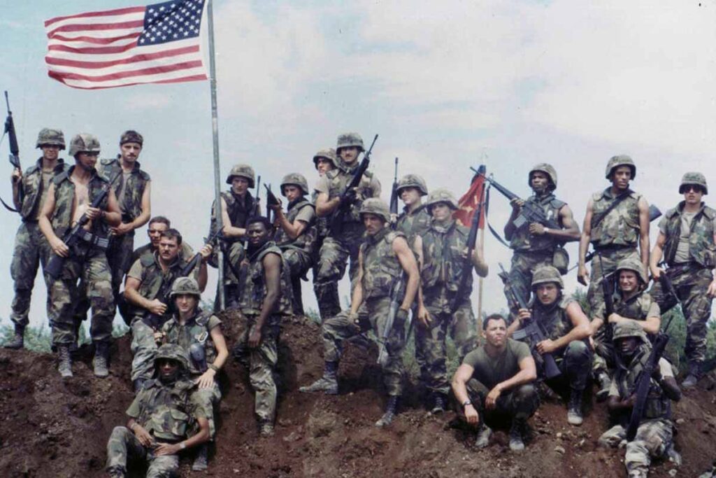 A group of U.S. soldiers in M81 Woodland BDU camouflage uniforms posing with an American flag, showing how all four branches once wore the same camo.