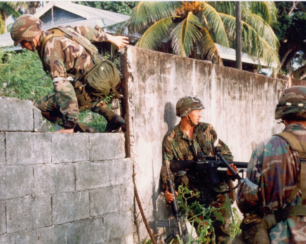 Three soldiers in M81 Woodland camouflage, one climbing over a wall and the other twoon guard below.