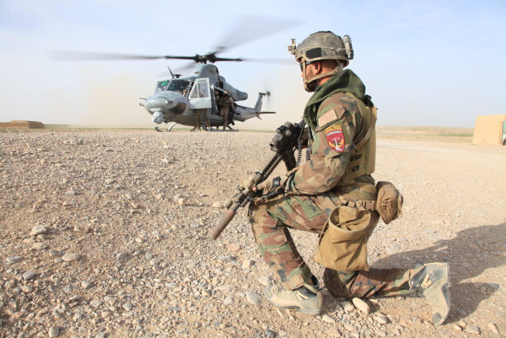 A Marine in U.S. Woodland camouflage kneels with a rifle as a helicopter lands in the background — an example of MARSOC’s return to Woodland during Afghanistan deployments. (U.S. Marine Corps photo by Cpl. Kyle McNally/Released)