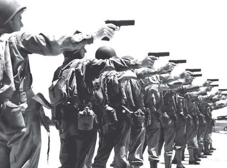 A black-and-white photo shows a line of soldiers in uniform holding pistols, all pointing them forward in a training exercise.