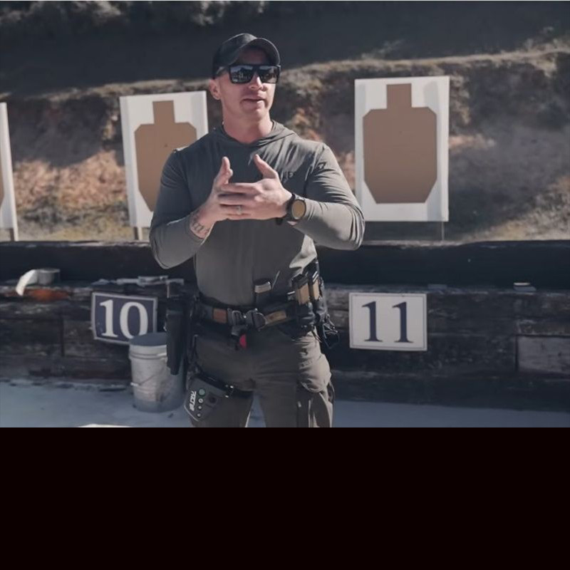 An instructor wearing a black hat and green shirt, holds his hands together as he talks to students about proper grip and how the thumbs are weak. 
