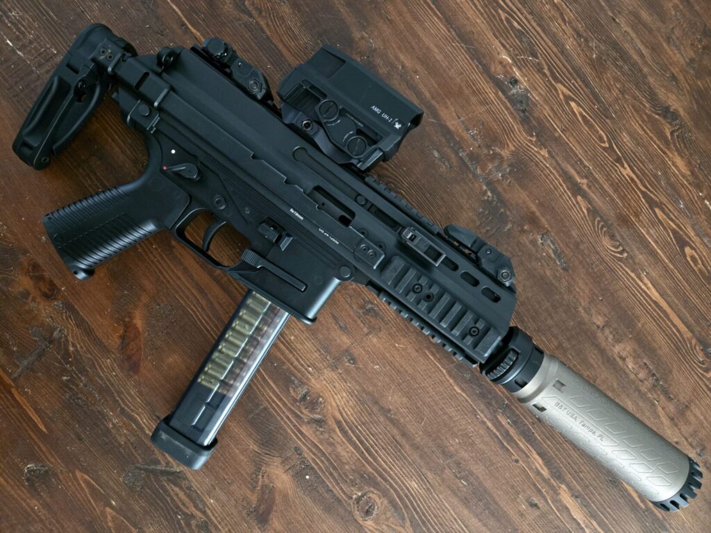 A firearm on a wooden backdrop with a holographic optic and suppressor