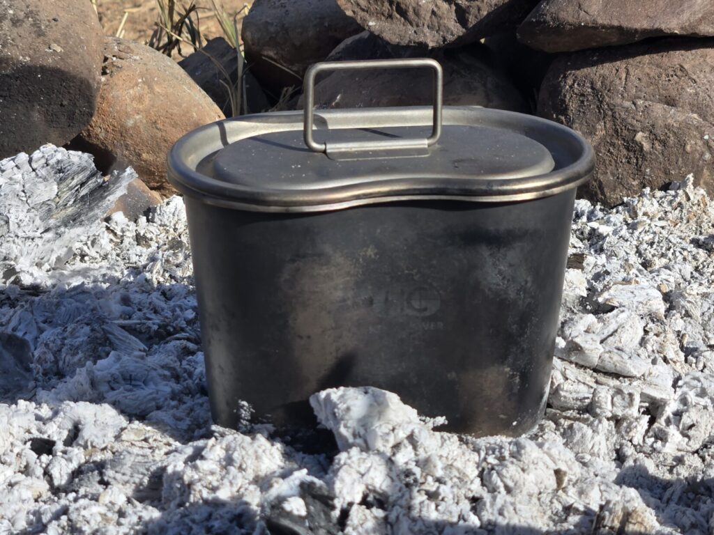 A titanium canteen cup with lid sitting in the ashes of a campfire