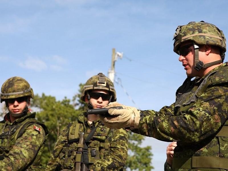 A person in military gear and camo clothes holds a Hi-Power pistol with both hands as he demonstrates something to other soldiers. 