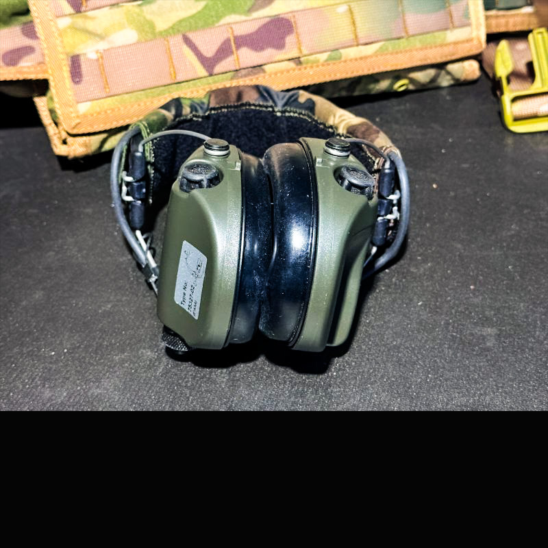 A pair of green earmuffs on a black table with a camo vest behind them.
