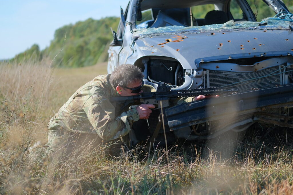 man fires a rifle from behind the cover provided by the front of a car