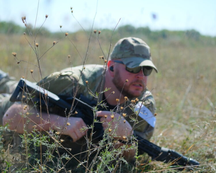 competitor low crawls with his rifle through brush