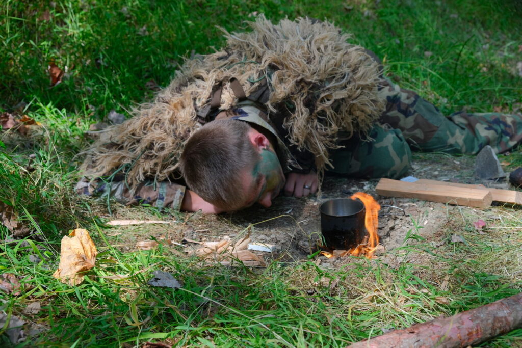 man in a ghillie suit blows on flames under a metal cup to boil water
