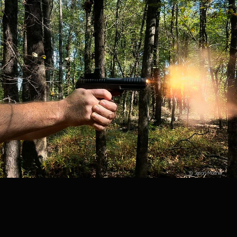 A view from the side of a persons arms and hands, shooting a PR57 handgun. Flames are coming out of the end of the gun as it goes off. 